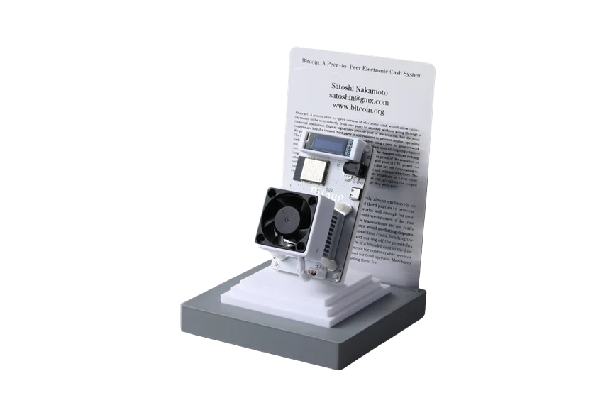 A Bitcoin mining rig on a stand, displayed with Satoshi Nakamoto's original Bitcoin whitepaper. The paper's title, "Bitcoin: A Peer-to-Peer Electronic Cash System," is visible.